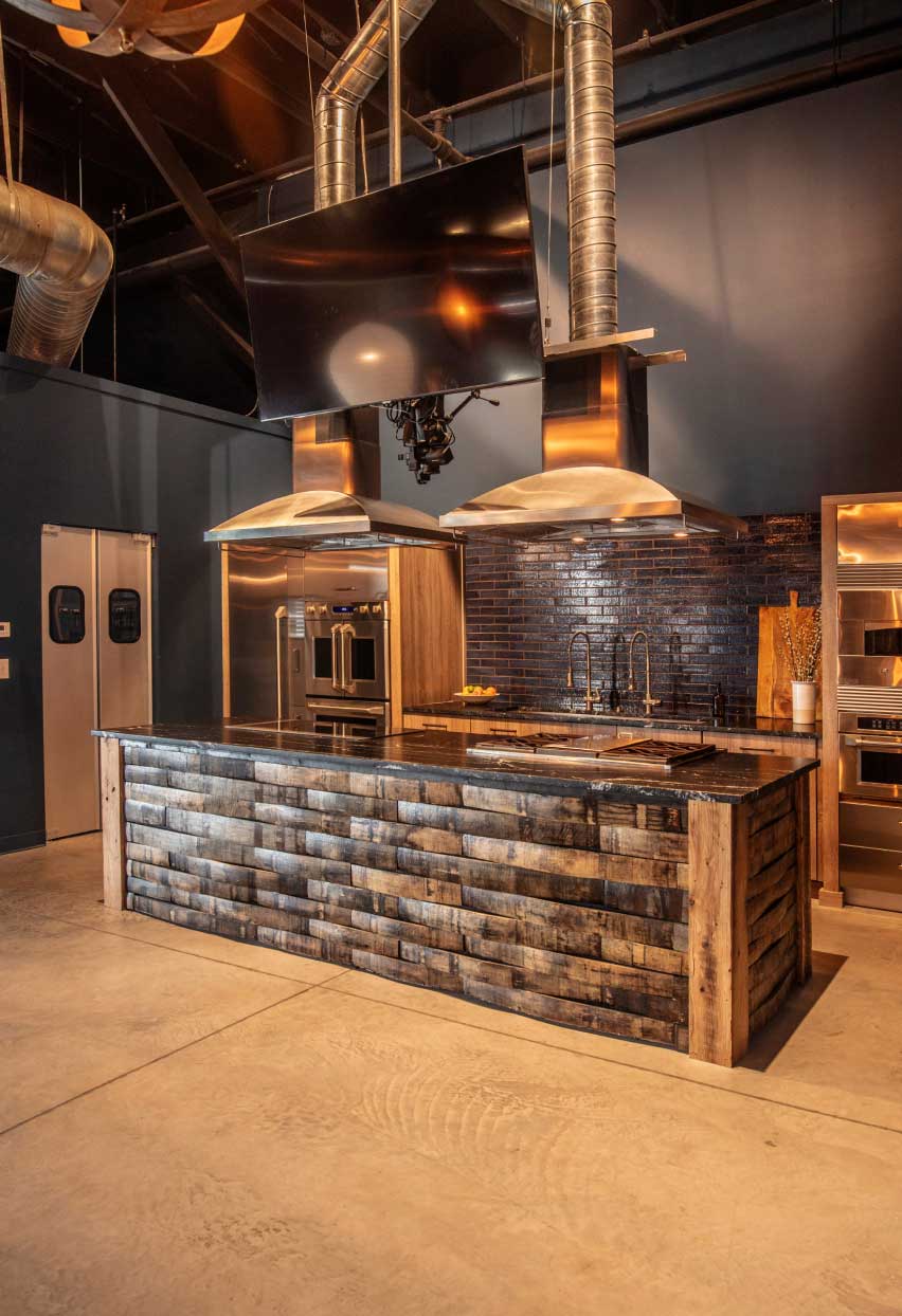 A modern rustic kitchen space at Bourbon Barrel Foods’ retail space with a cooktop, an oven and vent hoods
