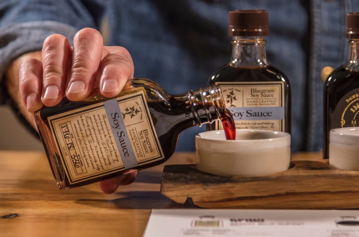A close-up of a hand pouring Bluegrass Soy Sauce into a small white bowl, with bottles of soy sauce in the background.