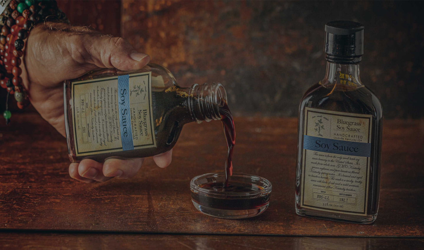 A hand holding a Bluegrass Soy Sauce bottle, pouring a serving into a dipping dish