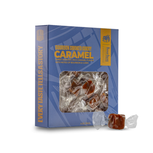 A blue box of Bourbon Smoked Honey Caramel candies with a clear window showing the wrapped candies inside, plus two loose wrapped candies placed in front on a white background.