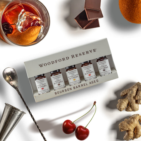A boxed set of four Woodford Reserve bourbon barrel aged whiskey distillate bottles arranged on a white surface with styled ingredients around it, including a drink with orange and ice, chocolate, an orange slice, ginger root, and two red cherries.