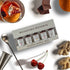 A boxed set of four Woodford Reserve bourbon barrel aged whiskey distillate bottles arranged on a white surface with styled ingredients around it, including a drink with orange and ice, chocolate, an orange slice, ginger root, and two red cherries.