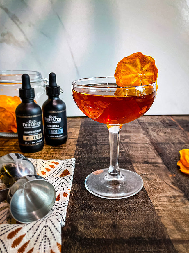 A craft cocktail served in an elegant coupe glass, garnished with a dried persimmon slice, with bottles of Old Forester bitters and bartending tools in the background.