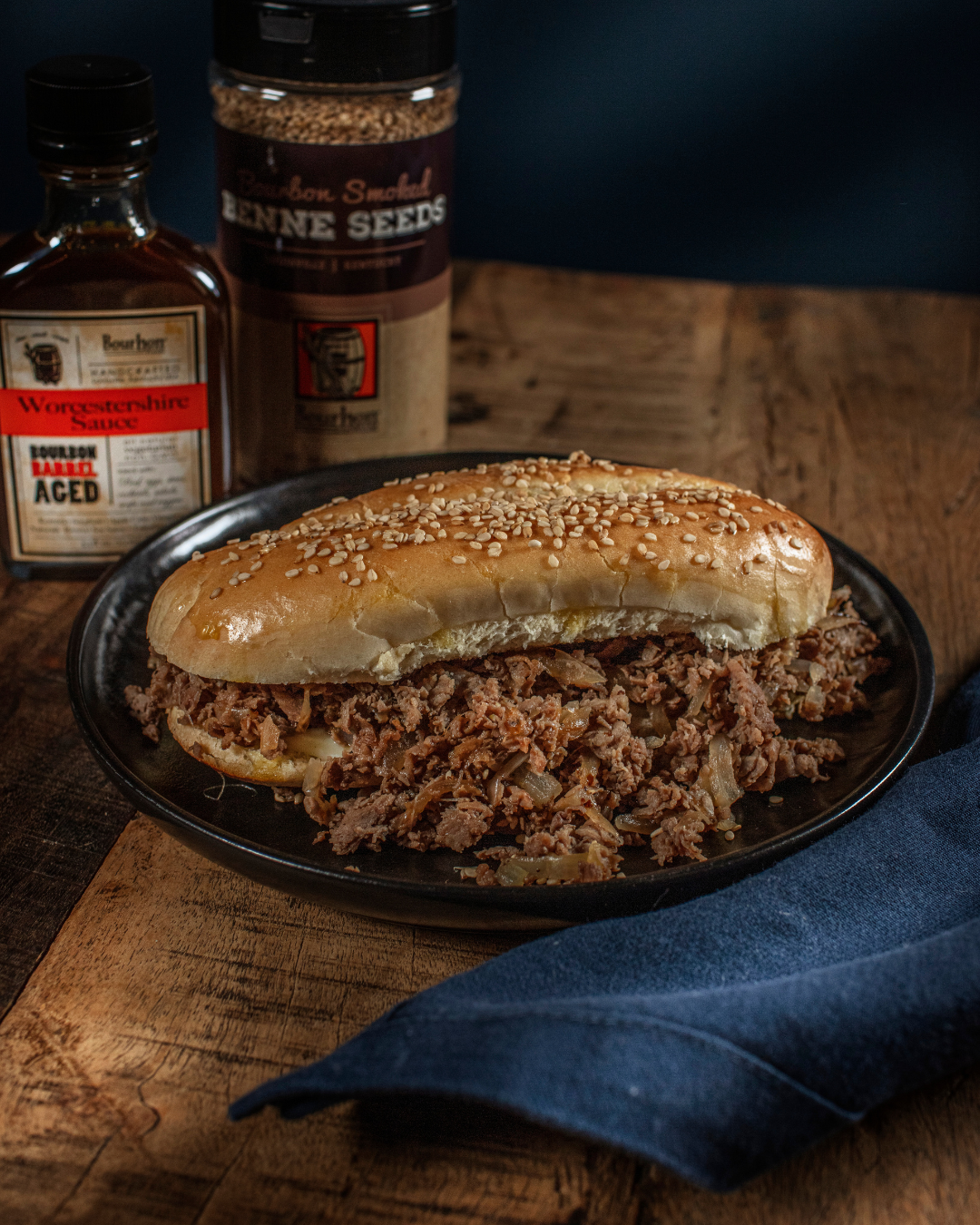 A sesame seed bun sandwich filled with seasoned shredded beef and onions, served on a black plate with Worcestershire sauce nearby.