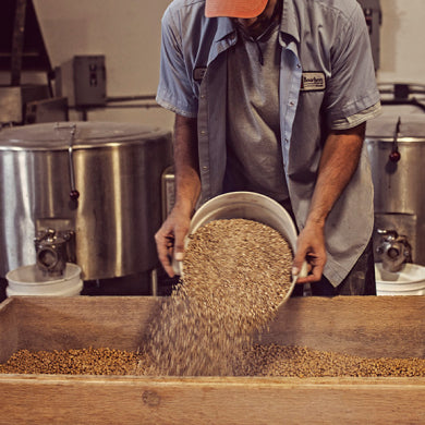A man using a bucket to pour a soybean and wheat mixture into a wooden tray