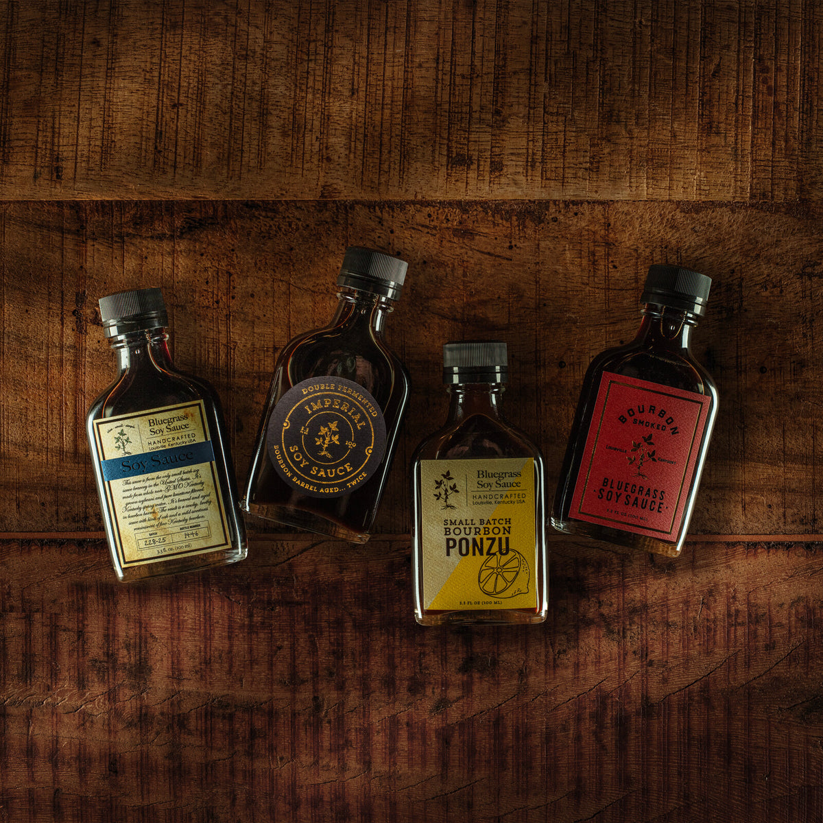 Four Bourbon Barrel Foods bottles — Bluegrass Soy Sauce, Imperial Double Fermented Soy Sauce, Small Batch Bourbon Ponzu and Bluegrass Soy Sauce — laid out on a dark wooden surface