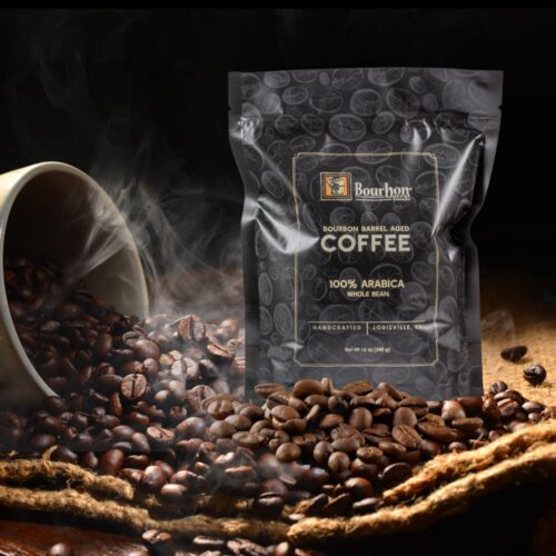 Bourbon Barrel Aged Coffee - one hundred percent Arabica whole bean coffee. Shown in a twelve ounce bag handcrafted in Louisville, Kentucky. Lifestyle shot with Bourbon Barrel Aged whole coffee grounds and smoke.